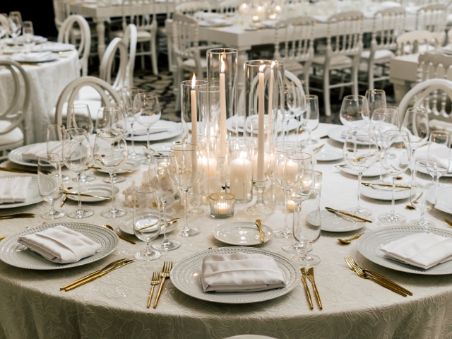 White party table setting with white tablecloth, white plates, gold silverware, and white candles of various sizes.