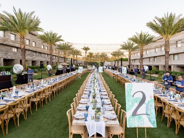 Full view of the king tables with wooden chairs, white table runners, blue glasses, and small cacti centerpieces including large table number signs.