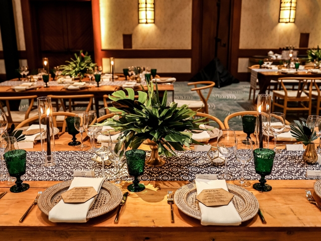 Place setting at awards dinner with wicker like charger plate, small wooden menu, green glasses, and foliage centerpiece.