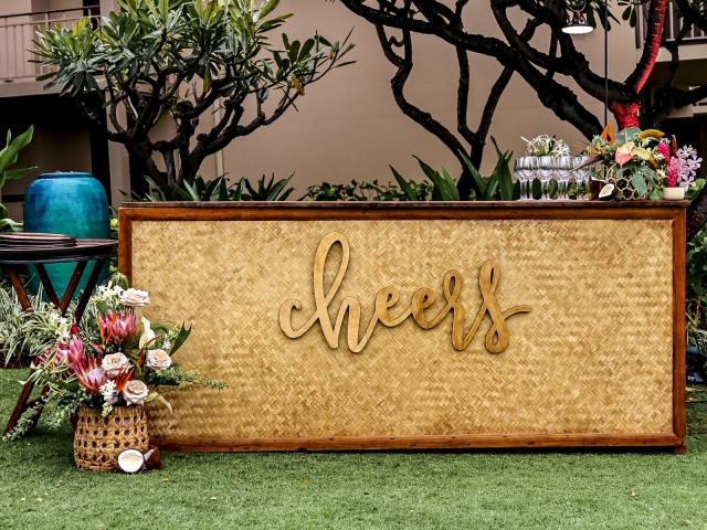 A bar with a golden cheers sign on the front with a floral display on the ground and on the bar.