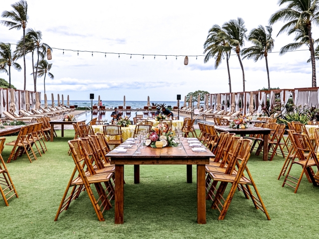 A full view of an outdoor welcome event in Hawaii with wooden tables overlooking the ocean.