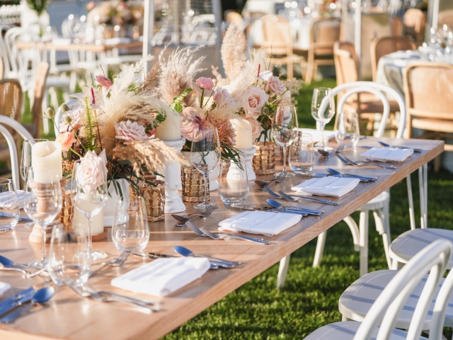 Long vintage  wooden table with white chairs and feathered floral arrangement along the center of the full table.