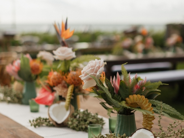 Large centerpieces for a long picnic-like table including an open coconut and tropical plants.