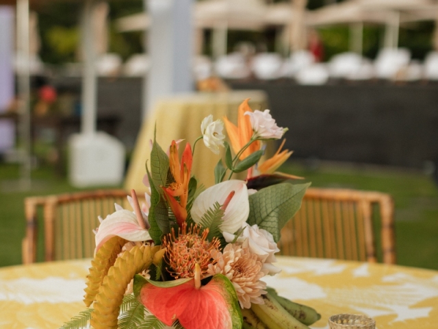 A close up of a tropical centerpiece including bright flowers and candles on a white and yellow tablecloth.