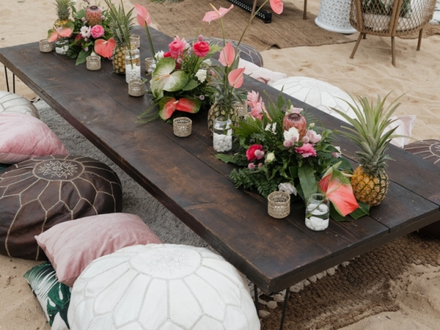 Low top long table on the beach with a floral centerpiece and cushion seats.