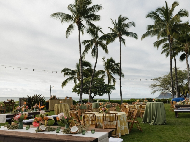 Outdoor event space with a variety of style tables, wooden chairs, overlooking the ocean with palm trees.