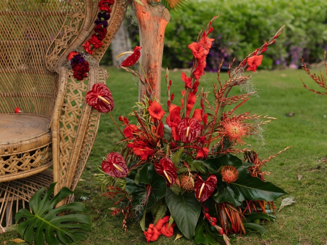 Red floral display arrangement sitting next to large wicker chair.