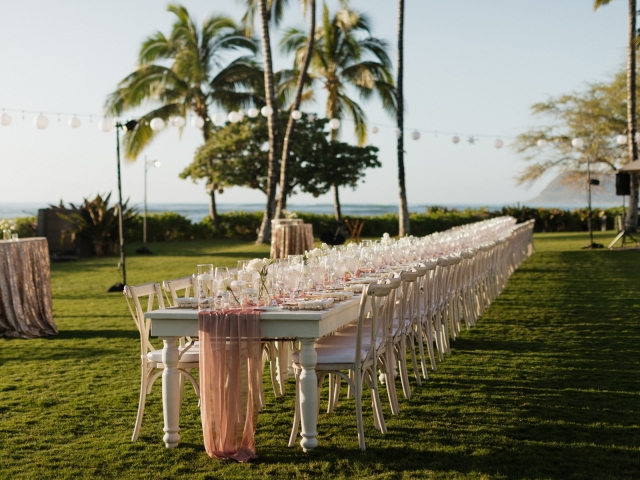 View of a long wooden table with champagne table runner, gold silverware, classic white floral centerpieces overlooking palm trees and water in Hawaii.